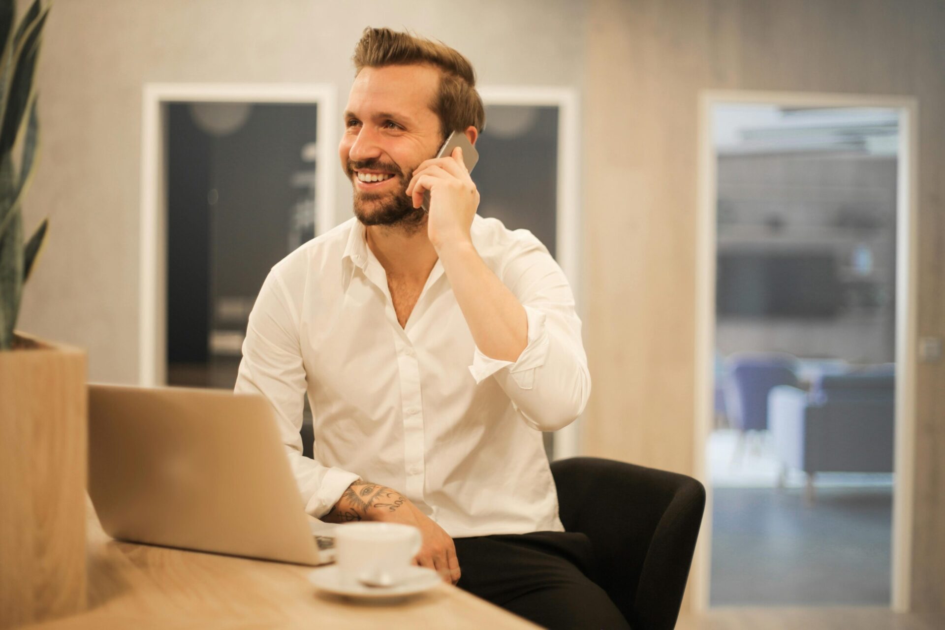 Happy businessman in white shirt on phone call at modern office desk with laptop.